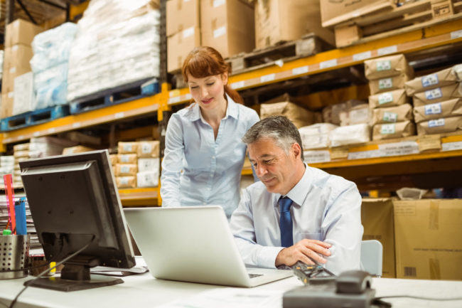 Warehouse management talking and looking at laptop in a large warehouse